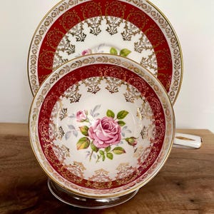 Pode incluir: Um conjunto de ch&aacute; vintage composto por uma ch&aacute;vena e um pires. A ch&aacute;vena e o pires apresentam um fundo branco com uma borda vermelha e dourada ornamentada. Uma rosa rosa est&aacute; centrada na ch&aacute;vena. O conjunto &eacute; exibido numa superf&iacute;cie de madeira.
