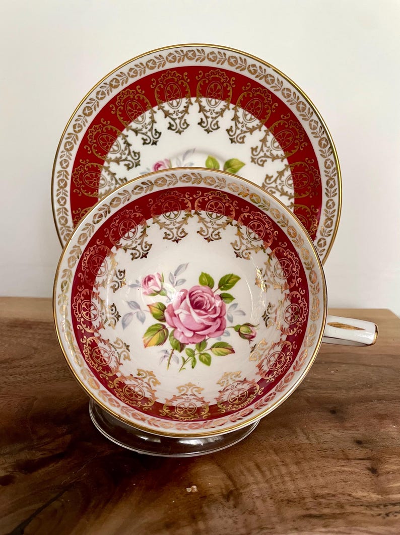 Pode incluir: Um conjunto de ch&aacute; vintage composto por uma ch&aacute;vena e um pires. A ch&aacute;vena e o pires apresentam um fundo branco com uma borda vermelha e dourada ornamentada. Uma rosa rosa est&aacute; centrada na ch&aacute;vena. O conjunto &eacute; exibido numa superf&iacute;cie de madeira.