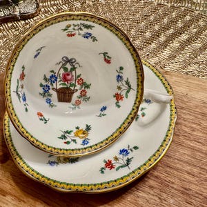 Pu&ograve; includere: Un set da t&egrave; bianco con bordo dorato e motivo floreale. La tazza presenta un cestino di fiori al centro, circondato da motivi floreali pi&ugrave; piccoli. Il piattino ha un motivo floreale simile.