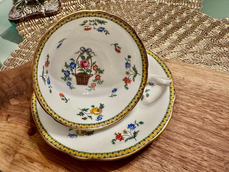 Pu&ograve; includere: Un set da t&egrave; bianco con bordo dorato e motivo floreale. La tazza presenta un cestino di fiori al centro, circondato da motivi floreali pi&ugrave; piccoli. Il piattino ha un motivo floreale simile.