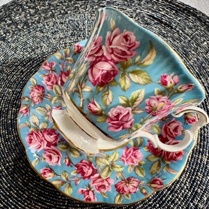 K&ouml;nnte beinhalten: Eine blaue und wei&szlig;e Teetasse und Untertasse mit einem floralen Muster aus rosa Rosen. Die Teetasse hat einen goldenen Rand und einen zarten Griff.