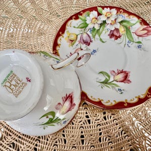 May include: A white teacup and saucer set with a floral design. The cup and saucer are decorated with red, pink, yellow, and green flowers. The rim of the saucer is red with a gold trim. The cup has a gold handle. The bottom of the cup reads "Narcissus Fine Bone China England".