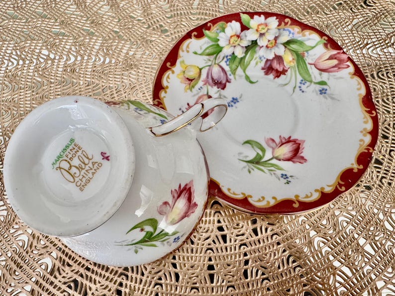 May include: A white teacup and saucer set with a floral design. The cup and saucer are decorated with red, pink, yellow, and green flowers. The rim of the saucer is red with a gold trim. The cup has a gold handle. The bottom of the cup reads "Narcissus Fine Bone China England".