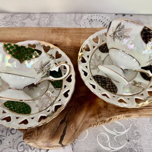Puede incluir: Dos tazas y platillos blancos con ribetes dorados y dise&ntilde;os de hojas. Los platillos tienen recortes en forma de coraz&oacute;n en los bordes. Las tazas y los platillos tienen acabados iridiscentes con acentos verdes y negros. Un juego de t&eacute; cl&aacute;sico.
