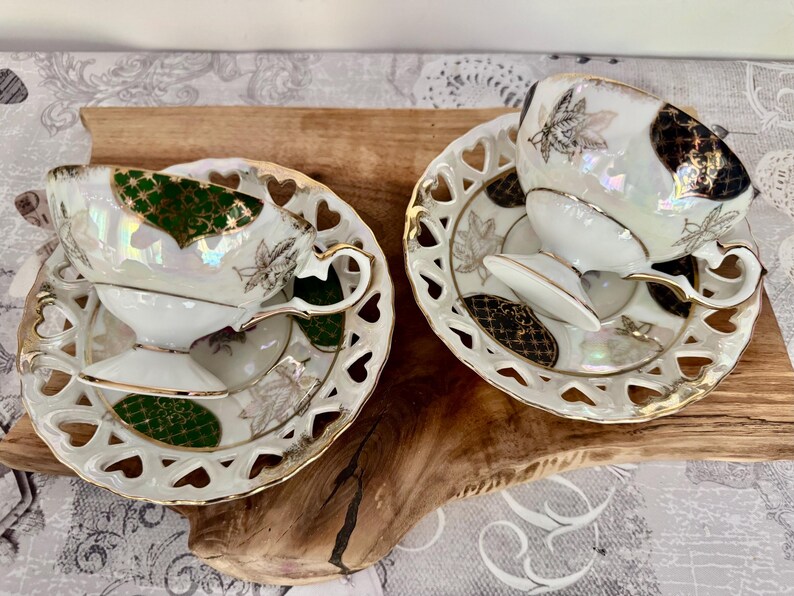 Puede incluir: Dos tazas y platillos blancos con ribetes dorados y dise&ntilde;os de hojas. Los platillos tienen recortes en forma de coraz&oacute;n en los bordes. Las tazas y los platillos tienen acabados iridiscentes con acentos verdes y negros. Un juego de t&eacute; cl&aacute;sico.