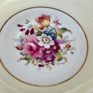 May include: A white saucer with a gold rim and a floral design in shades of pink, purple, blue, yellow, and green. The design features a large rose, smaller flowers, and leaves. The saucer is marked with the word "Fivonaro".