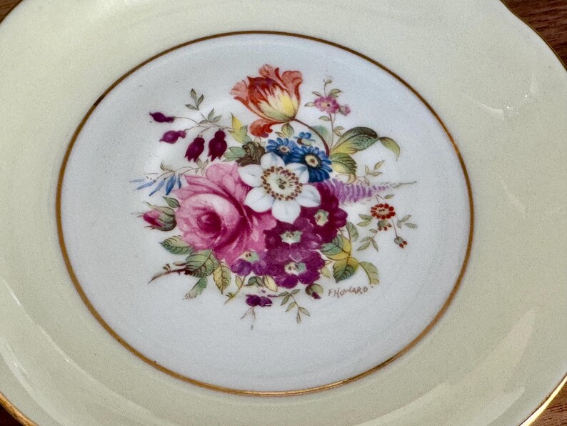 May include: A white saucer with a gold rim and a floral design in shades of pink, purple, blue, yellow, and green. The design features a large rose, smaller flowers, and leaves. The saucer is marked with the word "Fivonaro".