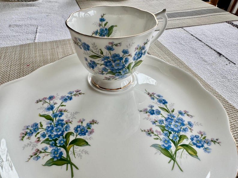 May include: A white porcelain teacup and saucer set with a blue and green floral pattern. The cup and saucer feature a delicate floral design with forget-me-not flowers.