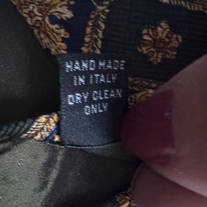May include: A dark green and navy blue necktie featuring a gold-toned ornate pattern. A black fabric tag is sewn onto the back, displaying the white text: HAND MADE IN ITALY DRY CLEAN ONLY. The tie fabric has a visible textured weave.