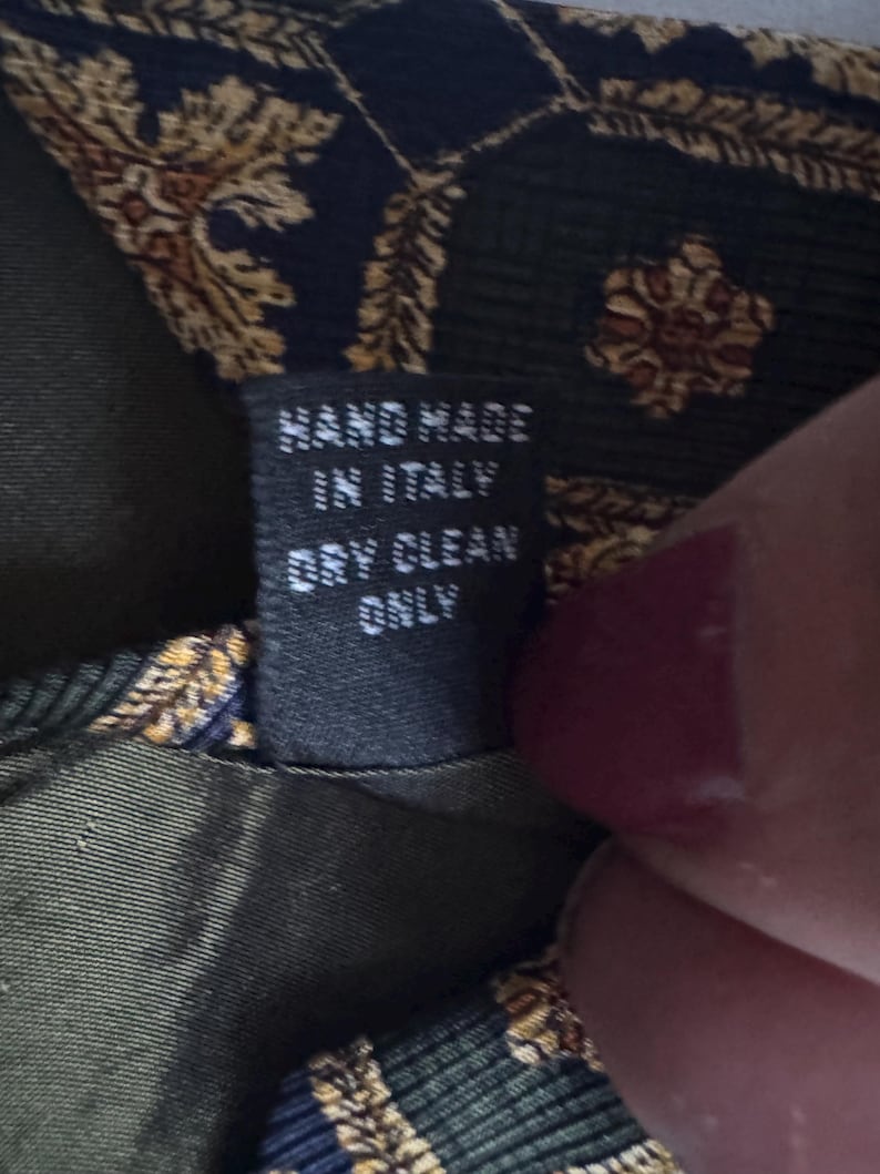 May include: A dark green and navy blue necktie featuring a gold-toned ornate pattern. A black fabric tag is sewn onto the back, displaying the white text: HAND MADE IN ITALY DRY CLEAN ONLY. The tie fabric has a visible textured weave.