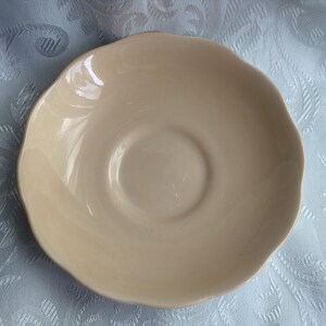 May include: A round, beige ceramic saucer with a smooth, glossy finish and a scalloped edge. The saucer features a central circular indentation designed to hold a cup, with a gently sloping rim that creates a shallow, basin-like profile.