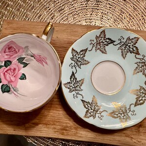 May include: A vintage teacup and saucer set with a floral design. The teacup has a pink background with pink roses and green leaves. The saucer has a light blue background with a gold leaf design.