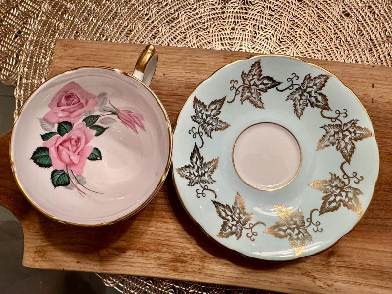 May include: A vintage teacup and saucer set with a floral design. The teacup has a pink background with pink roses and green leaves. The saucer has a light blue background with a gold leaf design.
