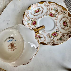 K&ouml;nnte beinhalten: Wei&szlig;es Tee-Set mit floralem Design und Goldverzierungen. Die Tasse hat einen goldenen Henkel. Sowohl die Tasse als auch die Untertasse sind mit rosa Rosen und gr&uuml;nen Bl&auml;ttern verziert. Auf der Unterseite der Tasse steht "Royal Albert Crown China England".