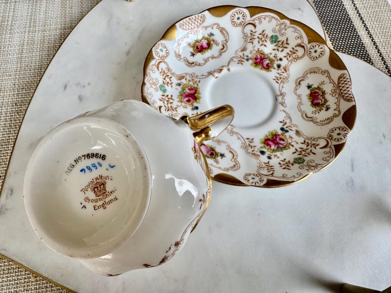 K&ouml;nnte beinhalten: Wei&szlig;es Tee-Set mit floralem Design und Goldverzierungen. Die Tasse hat einen goldenen Henkel. Sowohl die Tasse als auch die Untertasse sind mit rosa Rosen und gr&uuml;nen Bl&auml;ttern verziert. Auf der Unterseite der Tasse steht "Royal Albert Crown China England".