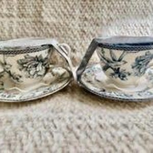 May include: Two ceramic teacups and matching saucers featuring a blue and white floral pattern. Each cup is secured with a white paper band wrapped around the top. The design includes intricate botanical illustrations and a decorative rim pattern.
