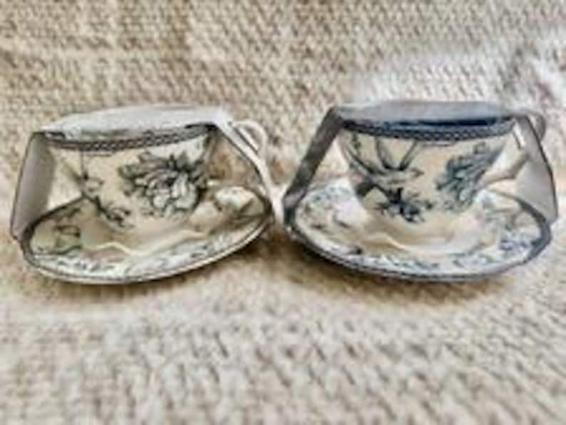 May include: Two ceramic teacups and matching saucers featuring a blue and white floral pattern. Each cup is secured with a white paper band wrapped around the top. The design includes intricate botanical illustrations and a decorative rim pattern.