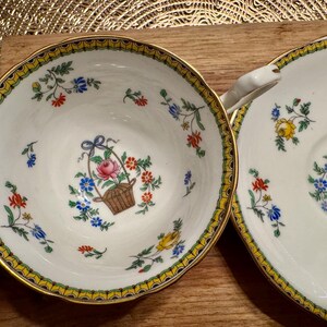 Pu&ograve; includere: Un set di due tazze da t&egrave; e piattini bianchi con un bordo giallo e dorato. Le tazze e i piattini presentano un motivo floreale con fiori rosa, gialli, blu e verdi. I fiori sono disposti in un cestino con un fiocco.