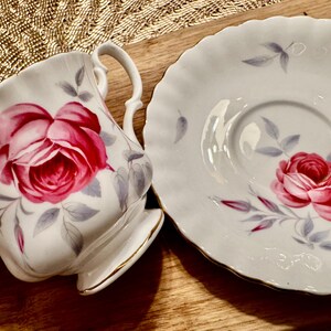 May include: A white teacup and saucer with a delicate floral design featuring pink roses and gray leaves. The cup and saucer have a scalloped edge.