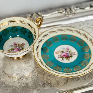 May include: A vintage teacup and saucer set. The teacup and saucer feature a teal and cream color scheme with gold accents. The center of the cup and saucer have a floral design. The teacup has a gold handle.
