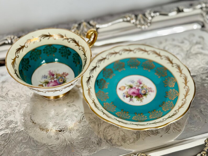 May include: A vintage teacup and saucer set. The teacup and saucer feature a teal and cream color scheme with gold accents. The center of the cup and saucer have a floral design. The teacup has a gold handle.