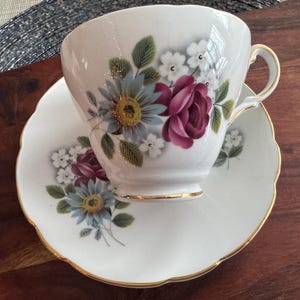 May include: A white teacup and saucer with a floral design. The design features pink, blue, and white flowers with green leaves. The cup and saucer have a gold trim.