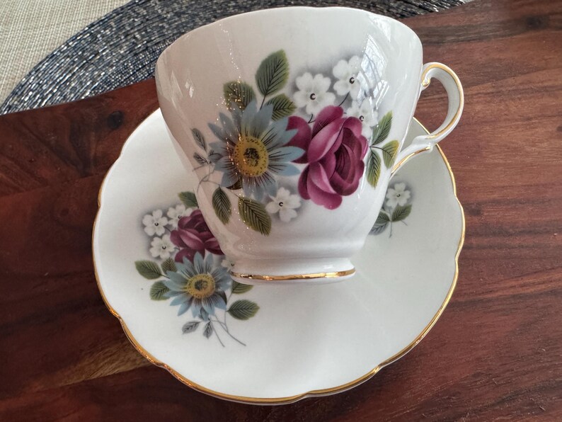 May include: A white teacup and saucer with a floral design. The design features pink, blue, and white flowers with green leaves. The cup and saucer have a gold trim.