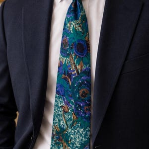 Pode incluir: Uma gravata com um padr&atilde;o floral vibrante em tons de azul-petr&oacute;leo, azul escuro, roxo e castanho, usada com uma camisa branca e um blazer azul-marinho. A gravata apresenta um design denso e pict&oacute;rico de grandes flores abstratas.