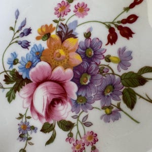 May include: Close-up of a white porcelain plate with a floral design. The design features a large pink rose, purple, blue, and yellow flowers, and green leaves. The plate has a gold rim.