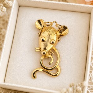 May include: A gold-colored mouse brooch with a whimsical design. The brooch features a mouse head with round eyes and large ears, wearing small glasses. The mouse's body is a curled tail. The brooch is presented in a white box.