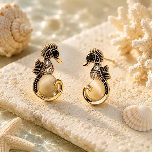 May include: A pair of gold-toned seahorse stud earrings with black and crystal accents, and a cream-colored pearl-like body. The earrings are displayed on a light-colored stone with seashells and coral in the background.