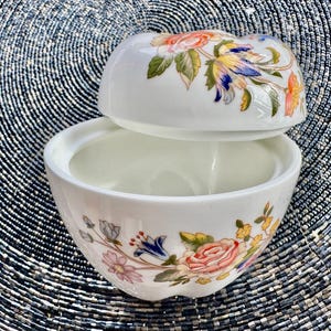 K&ouml;nnte beinhalten: Eine wei&szlig;e Porzellan-Schmuckdose mit Blumenmuster. Der Deckel ist ge&ouml;ffnet und die Dose ist leer. Die Dose ist mit bunten Blumen und Bl&auml;ttern verziert.