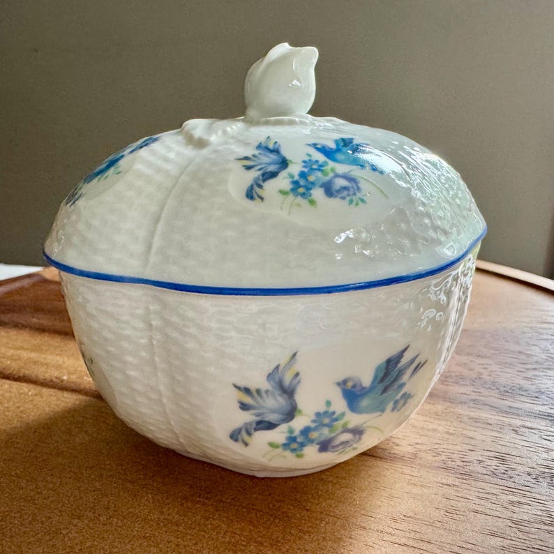 May include: White ceramic lidded dish with a blue floral and bird design. The dish has a woven texture and a blue trim. The lid is topped with a white flower-shaped handle. The design features blue birds and floral accents.