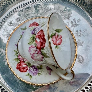 May include: A white teacup and saucer set with a floral design in red, pink, and purple, accented with gold trim. The teacup is resting on a matching saucer, set on a silver tray.