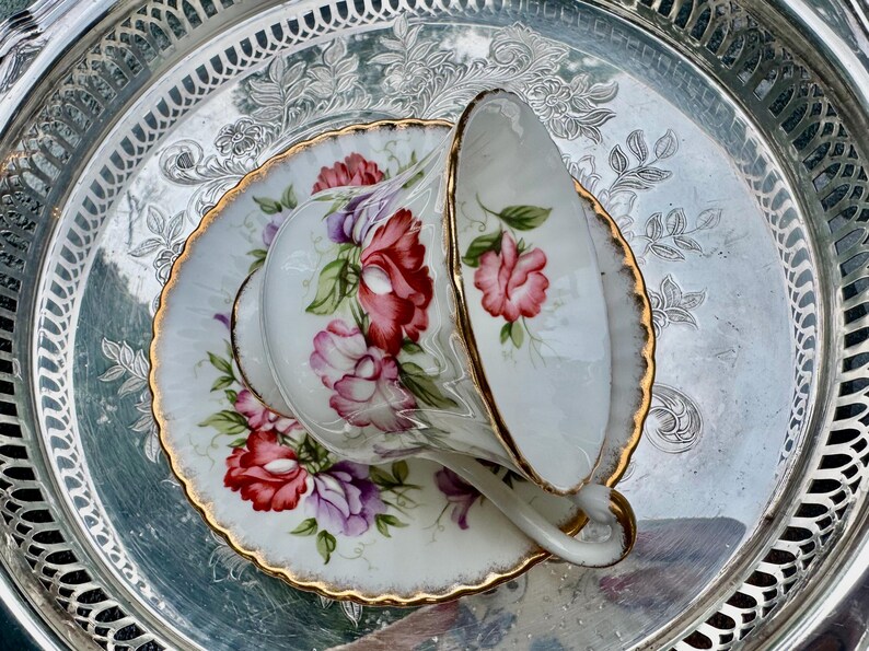 May include: A white teacup and saucer set with a floral design in red, pink, and purple, accented with gold trim. The teacup is resting on a matching saucer, set on a silver tray.