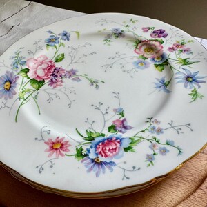 May include: A stack of white porcelain plates with a floral design. The plates have a gold rim and are decorated with pink, blue, purple, and yellow flowers and green leaves. The plates are stacked on a wooden serving tray.