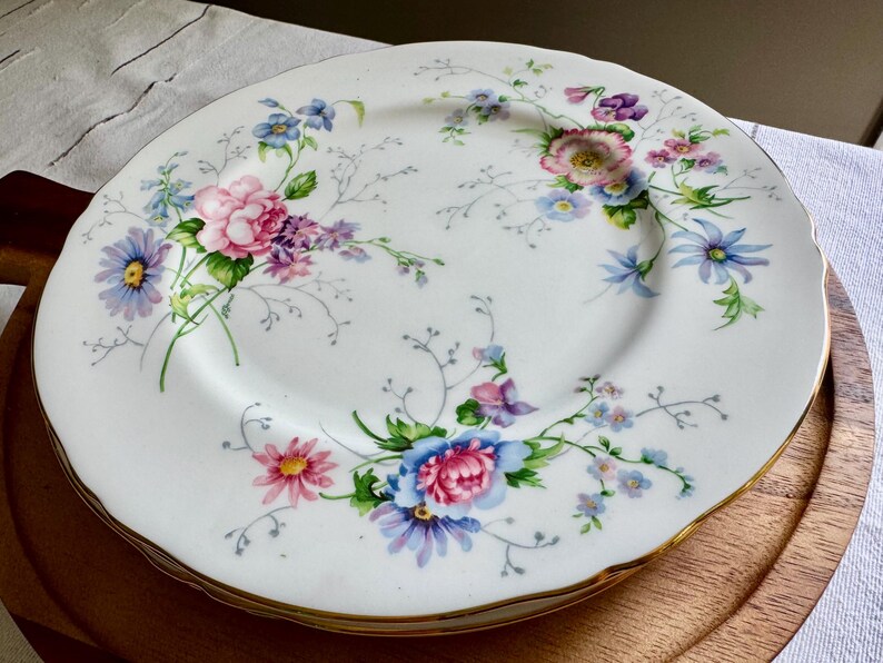 May include: A stack of white porcelain plates with a floral design. The plates have a gold rim and are decorated with pink, blue, purple, and yellow flowers and green leaves. The plates are stacked on a wooden serving tray.