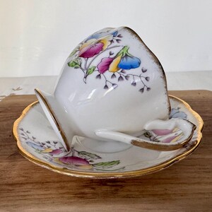 May include: A white teacup and saucer set with a floral design in pink, yellow, and blue. The cup is tilted, revealing the interior. The set has gold trim and is placed on a wooden surface. The handle of the cup is heart-shaped.