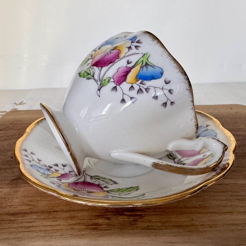 May include: A white teacup and saucer set with a floral design in pink, yellow, and blue. The cup is tilted, revealing the interior. The set has gold trim and is placed on a wooden surface. The handle of the cup is heart-shaped.