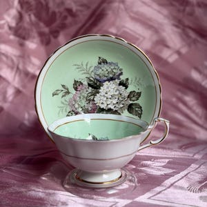 May include: A porcelain teacup and saucer set featuring a pale mint green interior and a floral motif of white and purple hydrangea blossoms. The edges of both the cup and saucer are trimmed with a thin gold band, and the cup has a pedestal base.