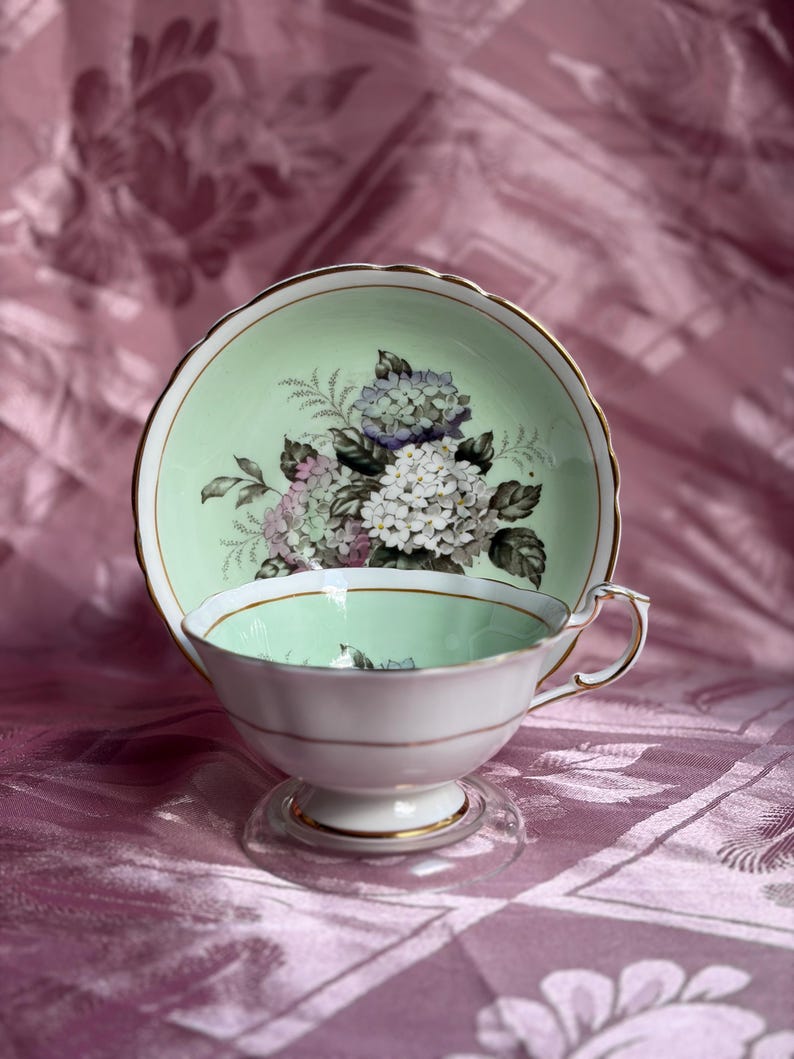 May include: A porcelain teacup and saucer set featuring a pale mint green interior and a floral motif of white and purple hydrangea blossoms. The edges of both the cup and saucer are trimmed with a thin gold band, and the cup has a pedestal base.