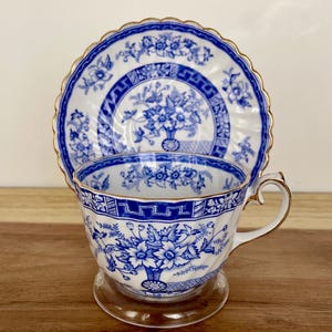 May include: A white teacup and saucer set with a blue floral design and gold trim. The teacup and saucer are displayed on a wooden surface. The teacup has a clear glass base.