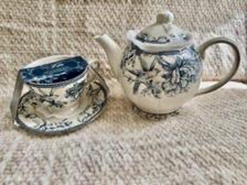 May include: A ceramic teapot and a matching teacup with a saucer, both featuring a blue and white floral pattern with bird illustrations. The teacup has a dark blue rectangular label attached to the rim. The set has a glossy finish.