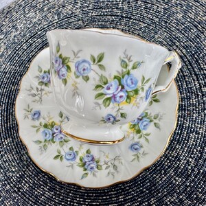 Pu&ograve; includere: Una tazza da t&egrave; e un piattino bianchi con un delicato motivo floreale con rose blu e foglie verdi. La tazza e il piattino hanno un bordo dorato.