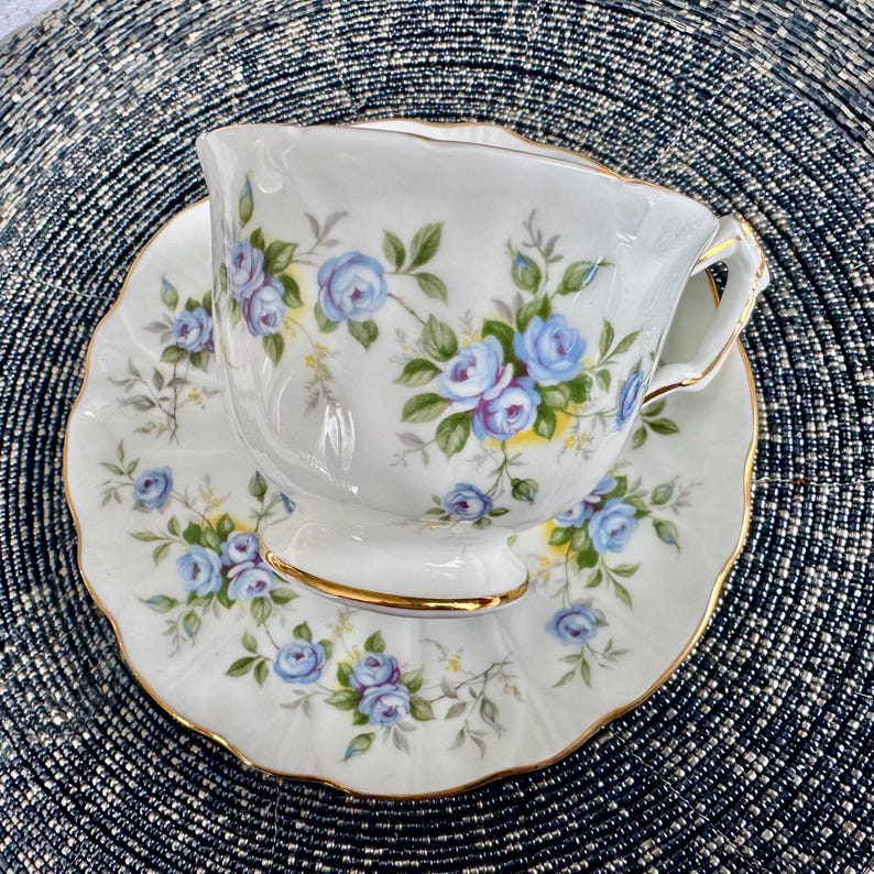 Pu&ograve; includere: Una tazza da t&egrave; e un piattino bianchi con un delicato motivo floreale con rose blu e foglie verdi. La tazza e il piattino hanno un bordo dorato.