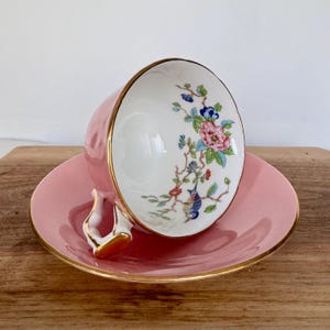 May include: Pink teacup and saucer set with gold trim. The teacup features a floral design with pink, blue, and green accents. The handle is white with gold detailing. The saucer is a matching pink color.