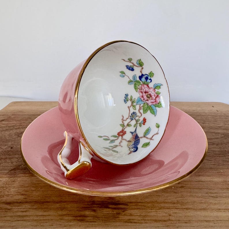 May include: Pink teacup and saucer set with gold trim. The teacup features a floral design with pink, blue, and green accents. The handle is white with gold detailing. The saucer is a matching pink color.