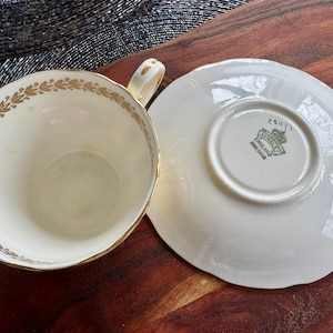 May include: A cream-colored bone china teacup and saucer set featuring a gold laurel leaf pattern along the inner rim of the cup. The saucer base displays a green stamp with the text EST 1775 AYNSLEY ENGLAND BONE CHINA and a crown icon.