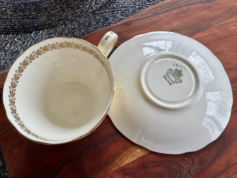 May include: A cream-colored bone china teacup and saucer set featuring a gold laurel leaf pattern along the inner rim of the cup. The saucer base displays a green stamp with the text EST 1775 AYNSLEY ENGLAND BONE CHINA and a crown icon.