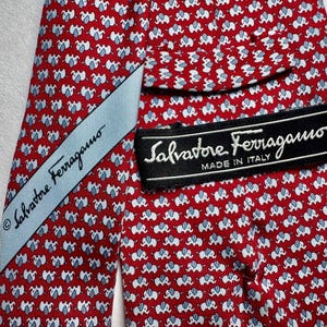 Puede incluir: Una corbata roja con un patr&oacute;n repetitivo de peque&ntilde;os elefantes blancos y azul claro. La corbata incluye una etiqueta de tela azul claro y una etiqueta negra con el texto Salvatore Ferragamo MADE IN ITALY impreso en blanco.